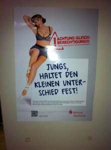 Ein weißes Plakat mit roter Schrift ACHTUNG GLEICHBERECHTIGUNG! und darunter in blau JUNGS HALTET DEN KLEINEN UNTERSCHIED FEST! Daneben eine weiße schlankge Frau in grauer Retro-Unterwäsche